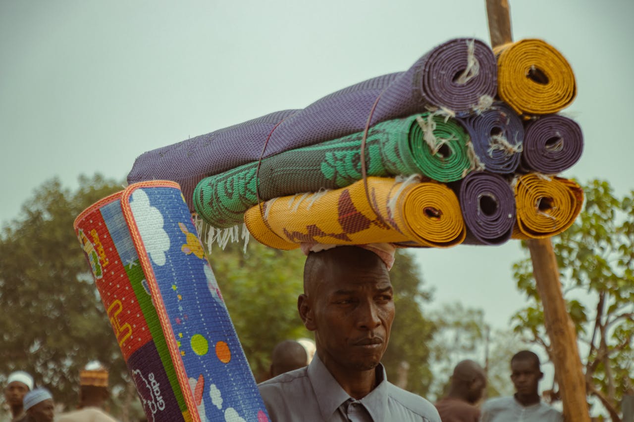 Man balancing colorful rolled mats on head at outdoor market. Summer.